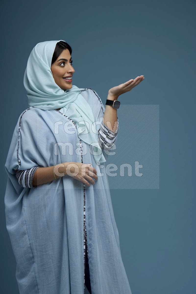 A Saudi woman pointing in a blue background wearing a Blue abaya