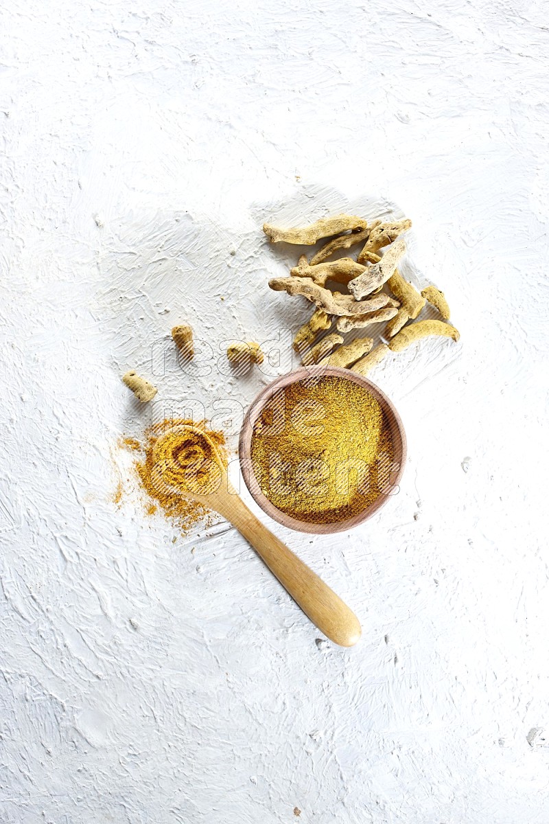 A wooden bowl and wooden spoon full of turmeric powder with dried turmeric fingers beside it on textured white flooring
