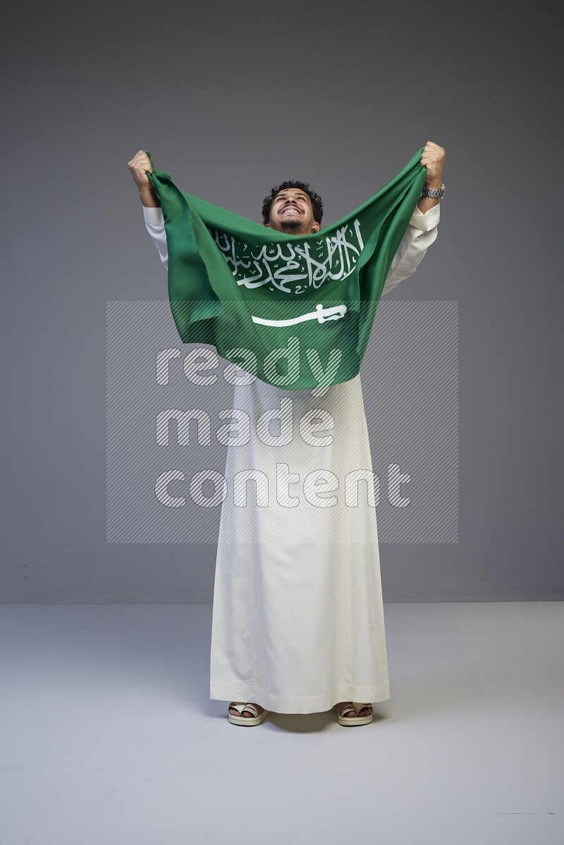 A Saudi man standing wearing thob holding big Saudi flag on gray background