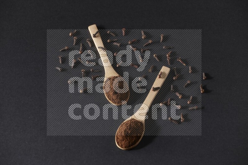 2 wooden spoons full of cloves powder with spreaded whole cloves on a black flooring