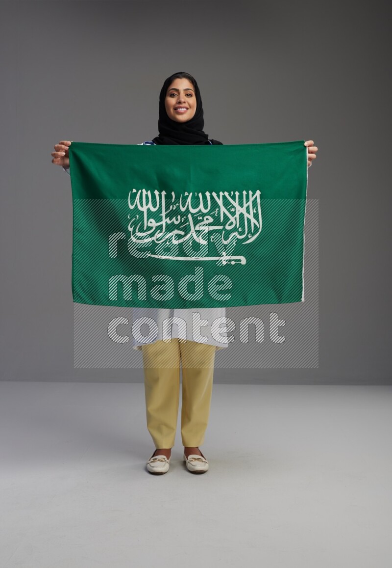 Saudi woman wearing lab coat with stethoscope standing holding Saudi flag on Gray background