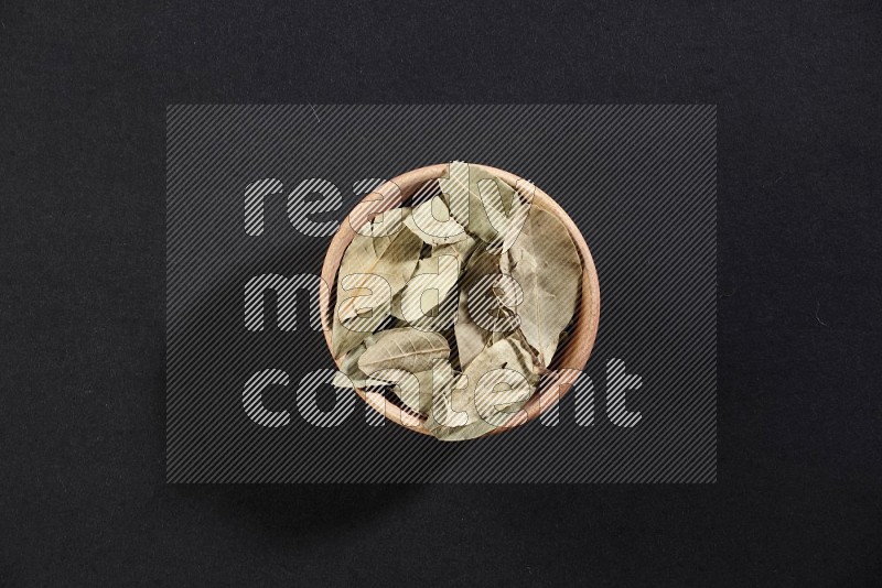 A wooden bowl filled with dried bay leaves on black flooring