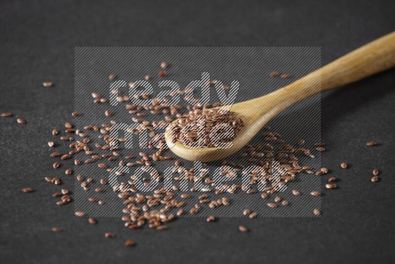 A wooden spoon full of flaxseeds and seeds spread beside it on a black flooring