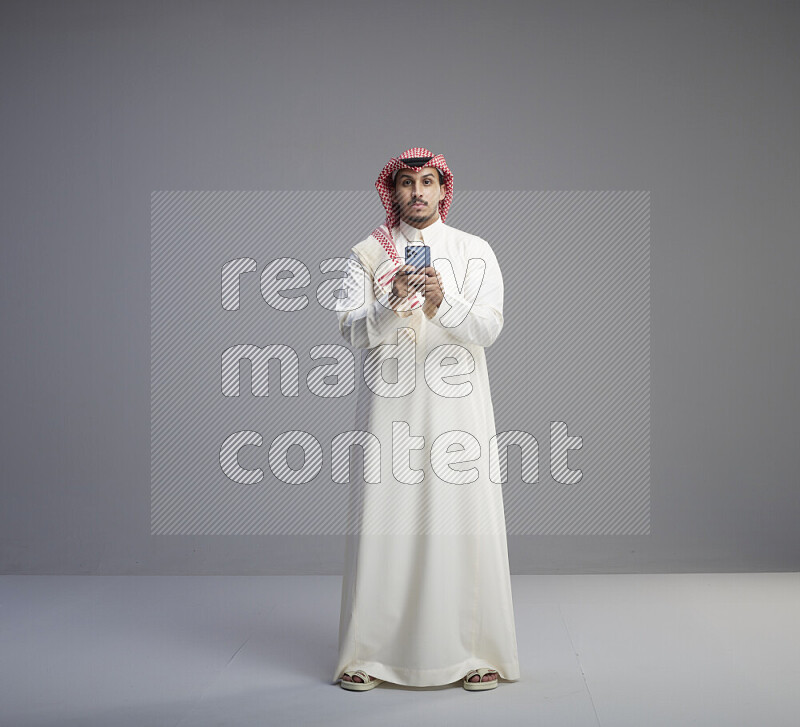 A  Saudi man standing wearing thob and red shomag texting on phone on gray background