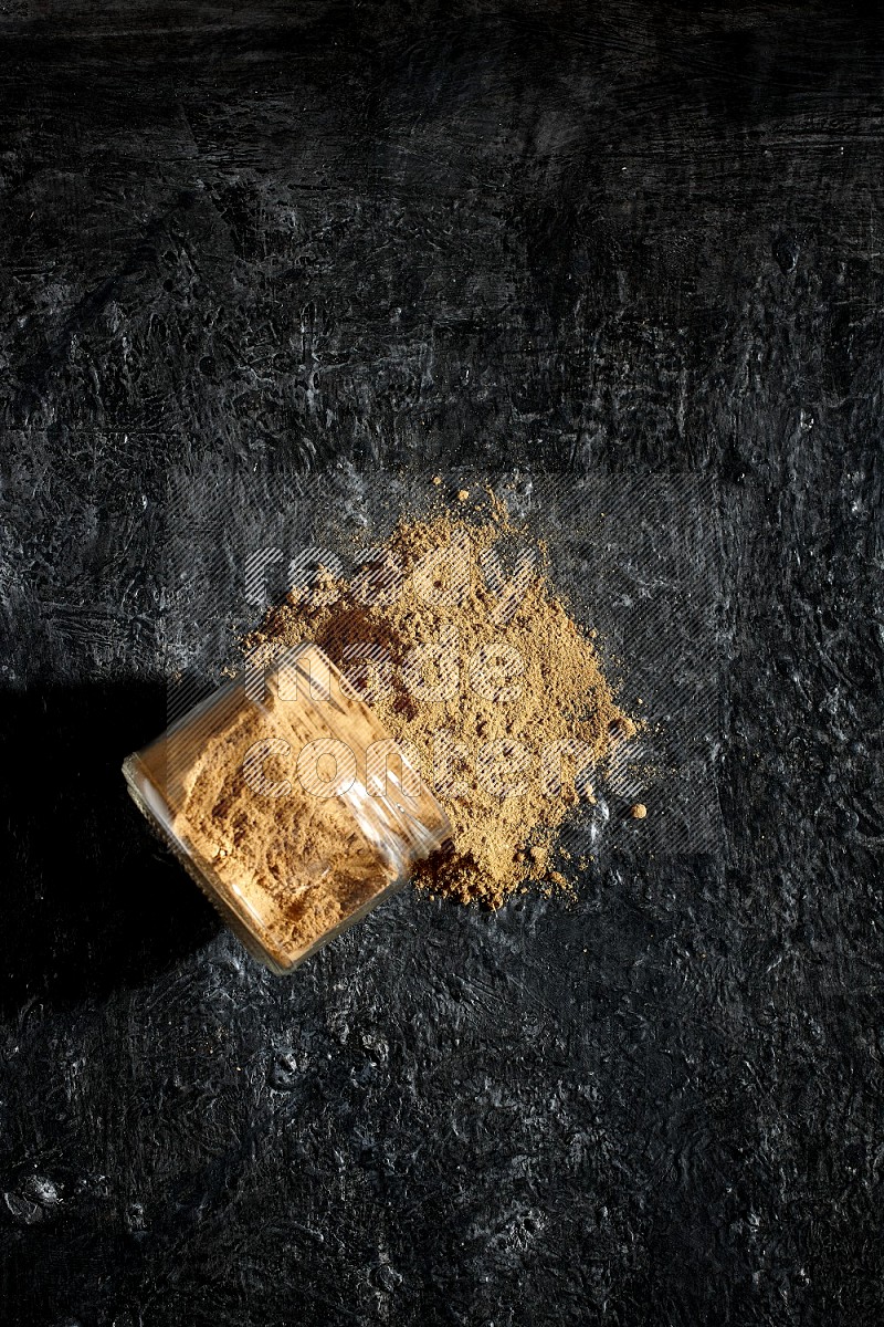 A flipped glass jar full of allspice powder and powder spilled out of it on a textured black flooring