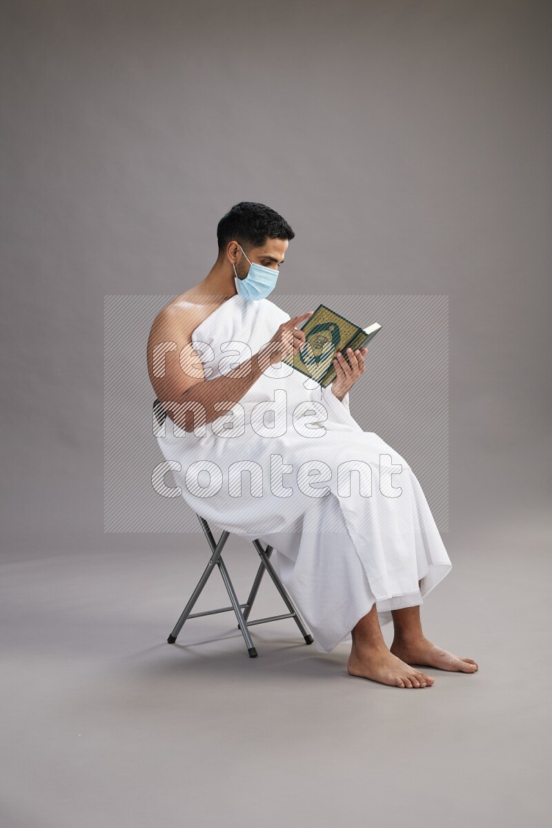 A man wearing Ehram with face mask sitting on chair reading quran on gray background
