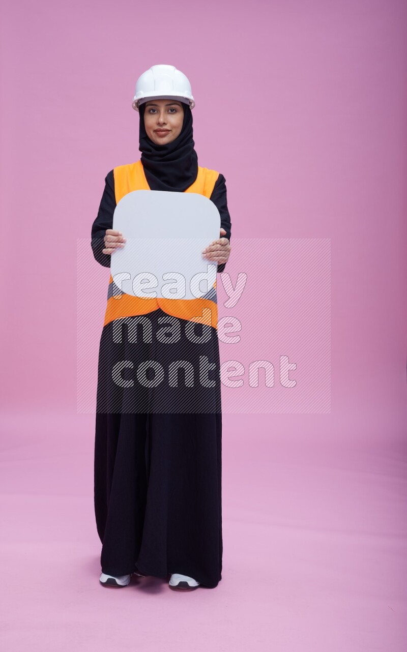 Saudi woman wearing Abaya with engineer vest and helmet standing holding social media sign on pink background