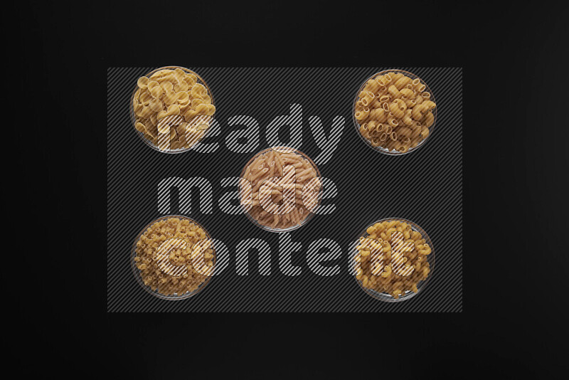 Different pasta types in 5 glass bowls on black background