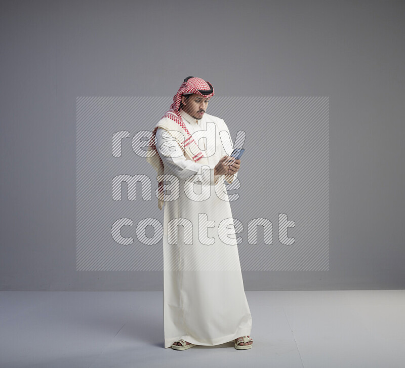 A  Saudi man standing wearing thob and red shomag texting on phone on gray background