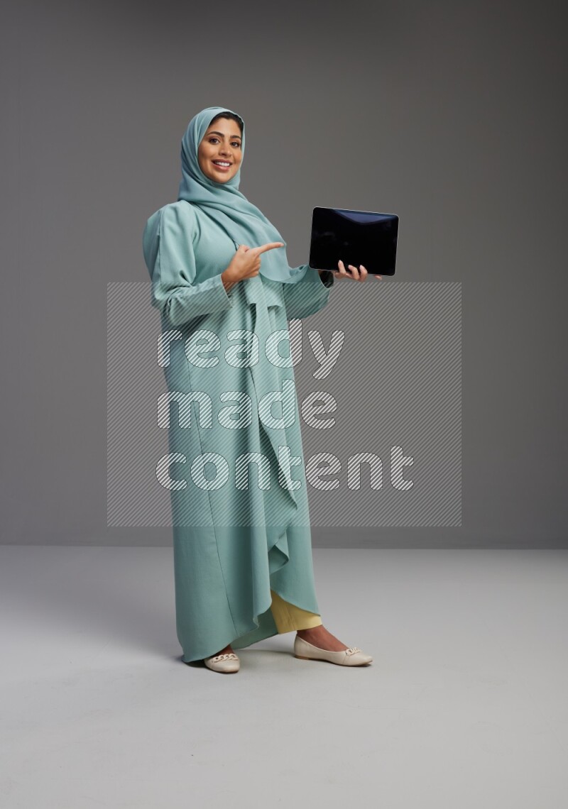 Saudi Woman wearing Abaya standing showing tablet to camera on Gray background