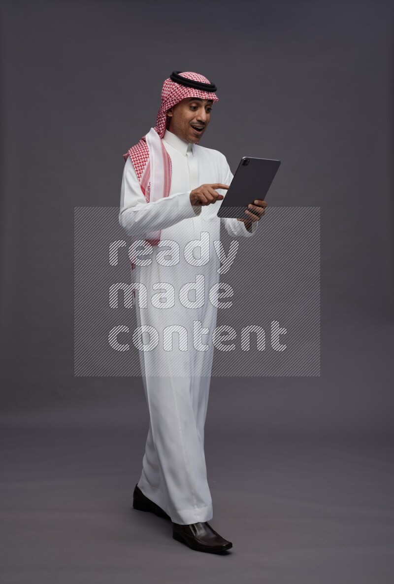 Saudi man wearing thob and shomag standing working on tablet on gray background