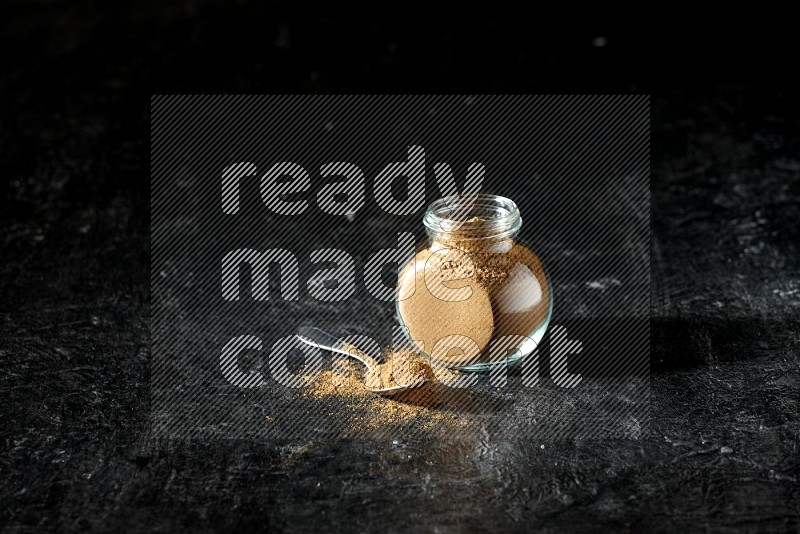 A glass spice jar and metal spoon full of allspice powder on a textured black flooring