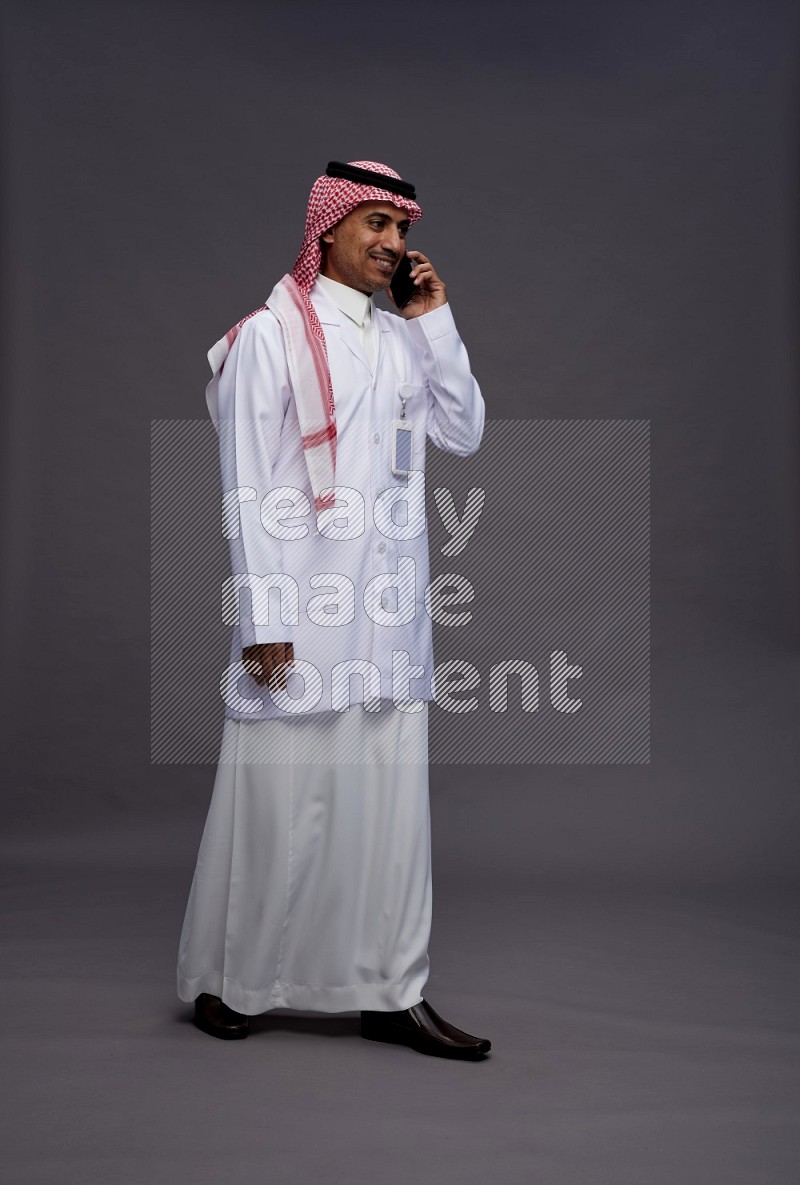 Saudi man wearing thob with lab coat and shomag with pocket employee badge standing talking on phone on gray background