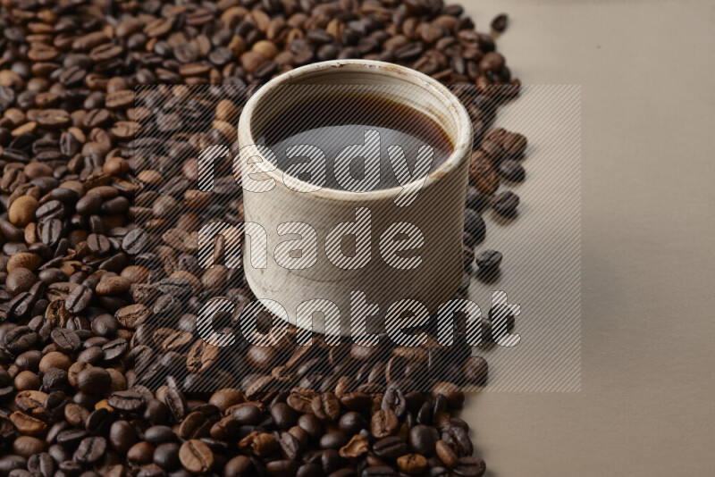 A beige pottery cup of coffee surrounded by roasted coffee beans on beige background