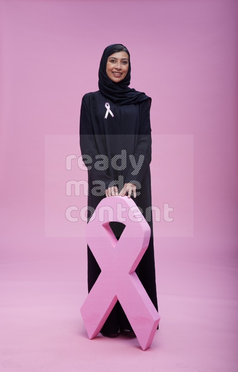 Saudi woman wearing pink ribbon on abaya standing holding awareness ribbon on plain pink background