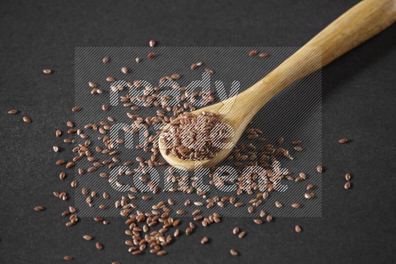 A wooden spoon full of flaxseeds and seeds spread beside it on a black flooring
