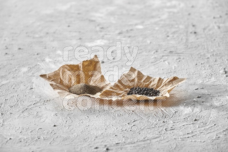 Black pepper and black pepper powder on 2 crumpled pieces of craft paper on a textured white flooring
