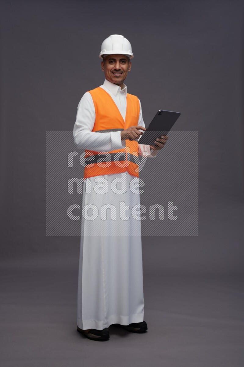 Saudi man wearing thob with engineer vest standing working on tablet on gray background