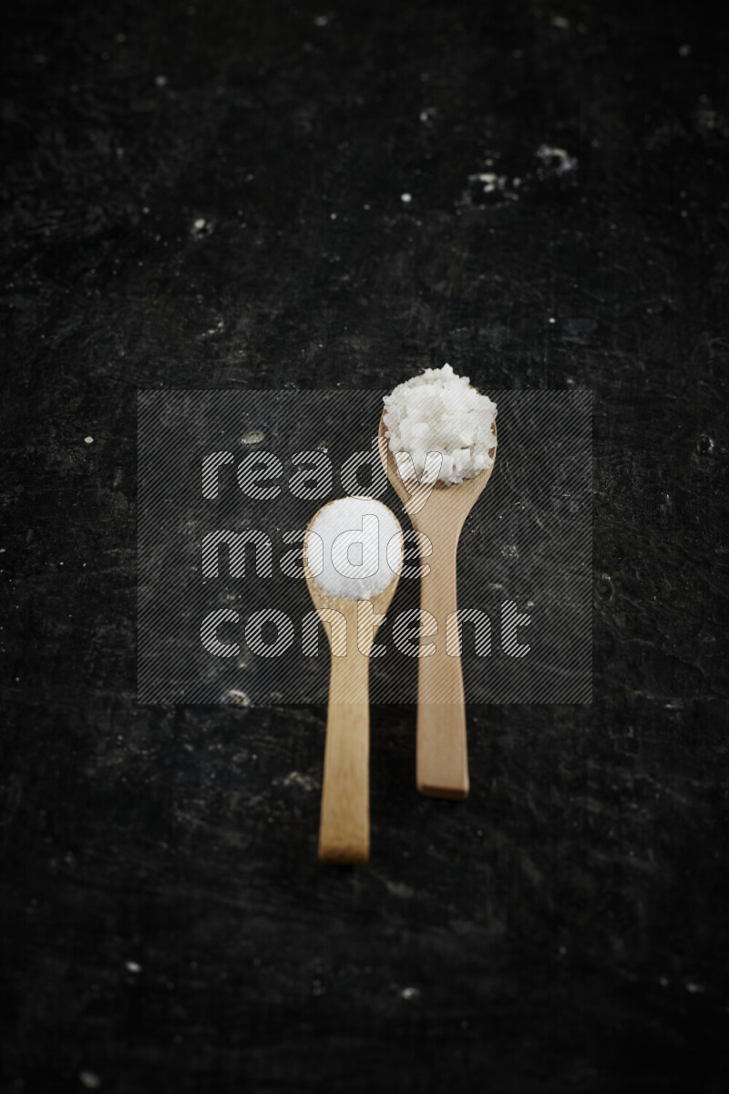 2 wooden spoons filled with fine and coarse salt on black background