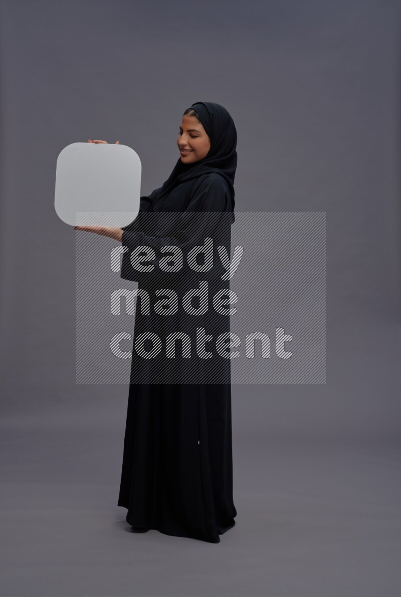 Saudi woman wearing Abaya standing holding social media sign on gray background