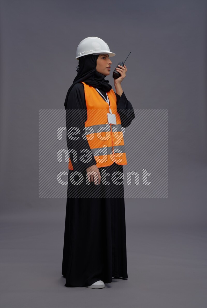 Saudi woman wearing Abaya with engineer vest with neck strap employee badge standing holding walkie-talkie on gray background