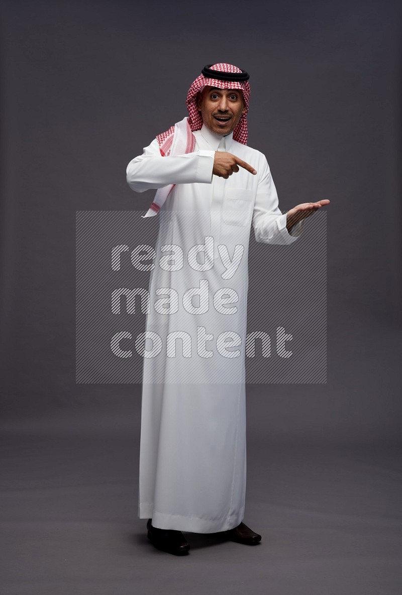 Saudi man wearing thob and shomag standing interacting with the camera on gray background