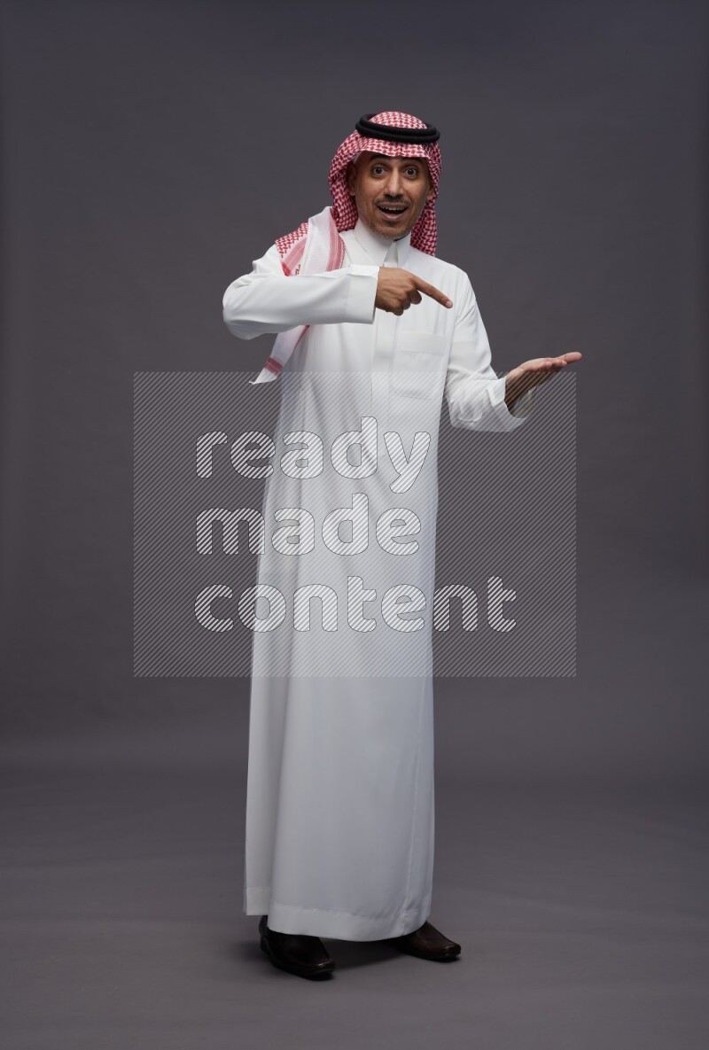 Saudi man wearing thob and shomag standing interacting with the camera on gray background