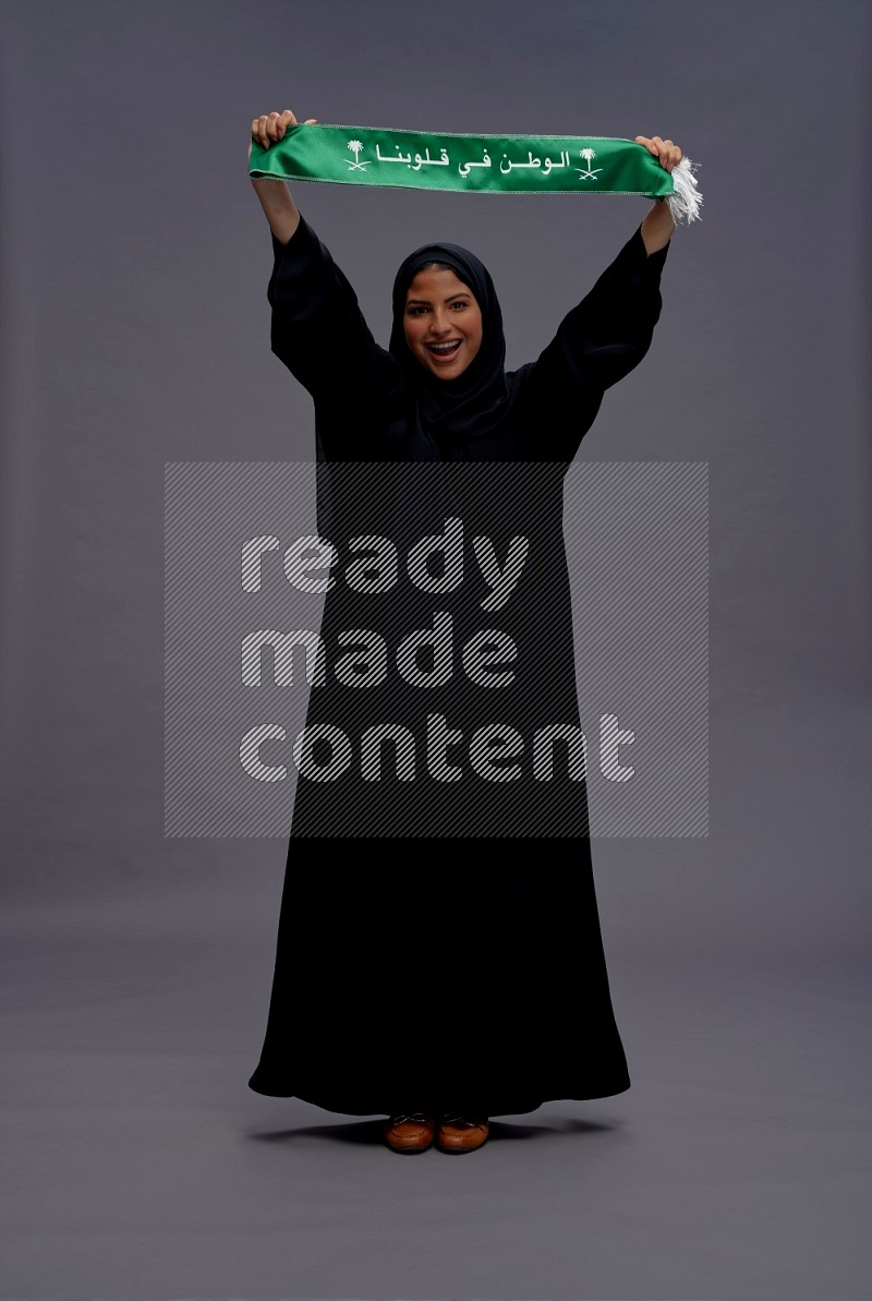 Saudi woman wearing Abaya standing holding Saudi flag on gray background