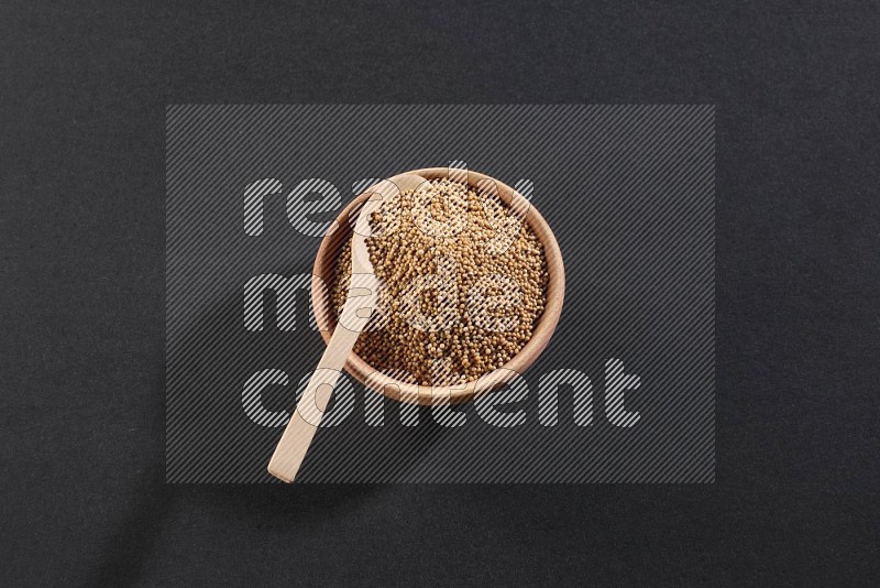 A wooden bowl and spoon full of mustard seeds on a black flooring