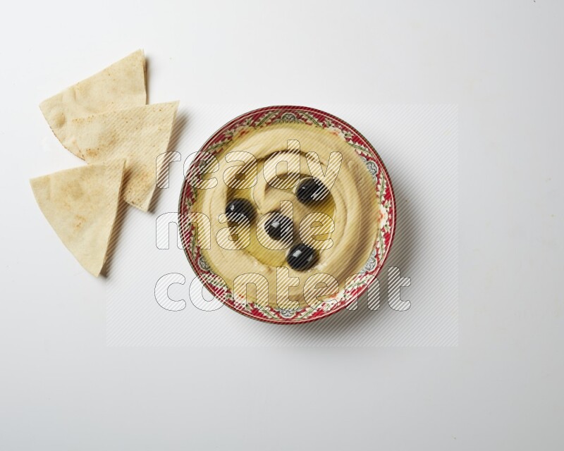 Hummus in a red plate with patterns garnished with black olives on a white background