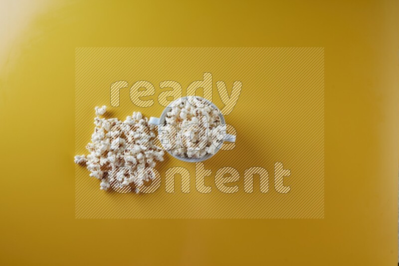 A white ceramic bowl full of popcorn with popcorn beside it on a yellow background in a top view shot