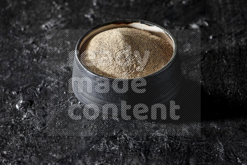 Black pottery bowl full of white pepper powder on textured black flooring