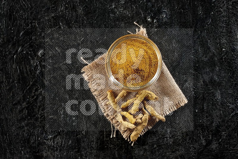 A glass bowl full of turmeric powder with dried turmeric fingers on a burlap fabric on textured black flooring