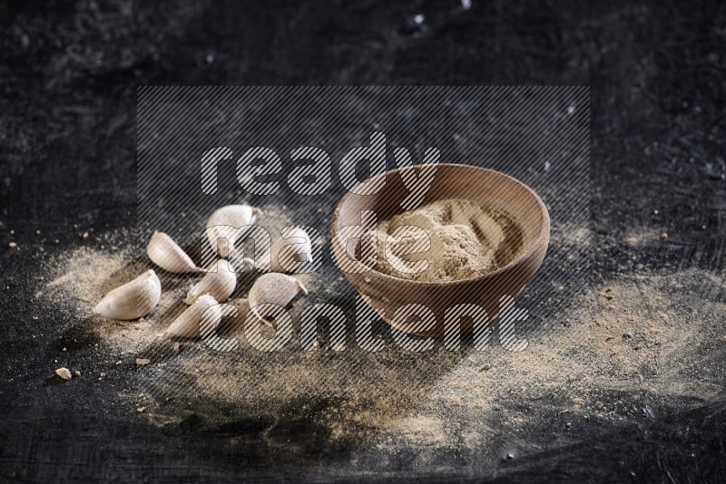 A wooden bowl full of garlic powder with some garlic cloves beside it on a textured black flooring