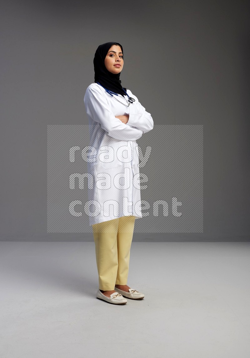 Saudi woman wearing lab coat with stethoscope standing with crossed arms on Gray background
