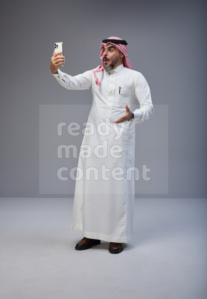 Saudi man Wearing Thob and red Shomag standing taking selfie on Gray background