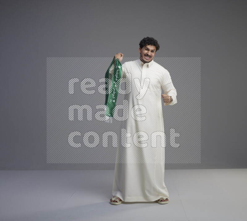 A Saudi man standing wearing thob holding Saudi flag scarf on gray background