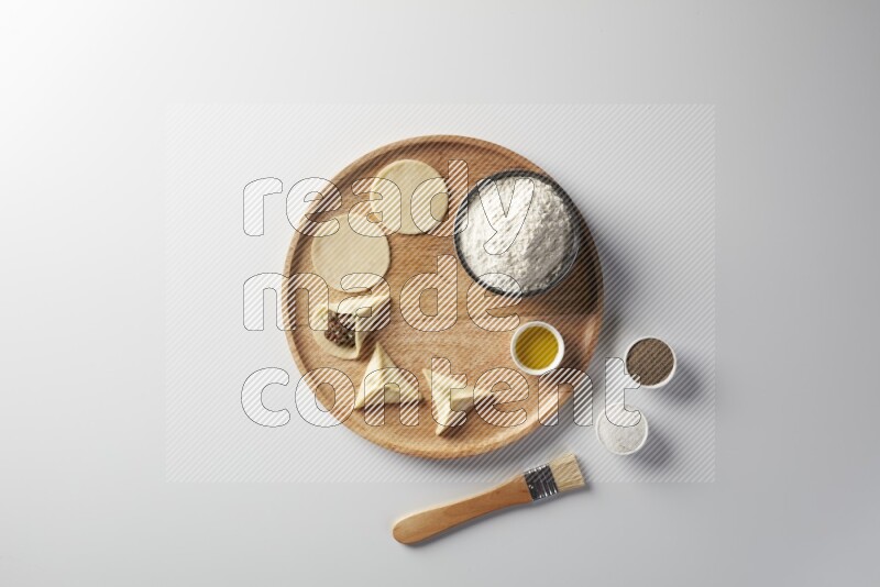 two closed sambosas and one open sambosa filled with meat while flour, salt, black pepper and oil with oil brush aside in a wooden dish on a white background