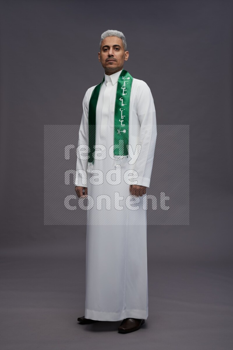 Saudi man wearing thob standing holding Saudi flag on gray background