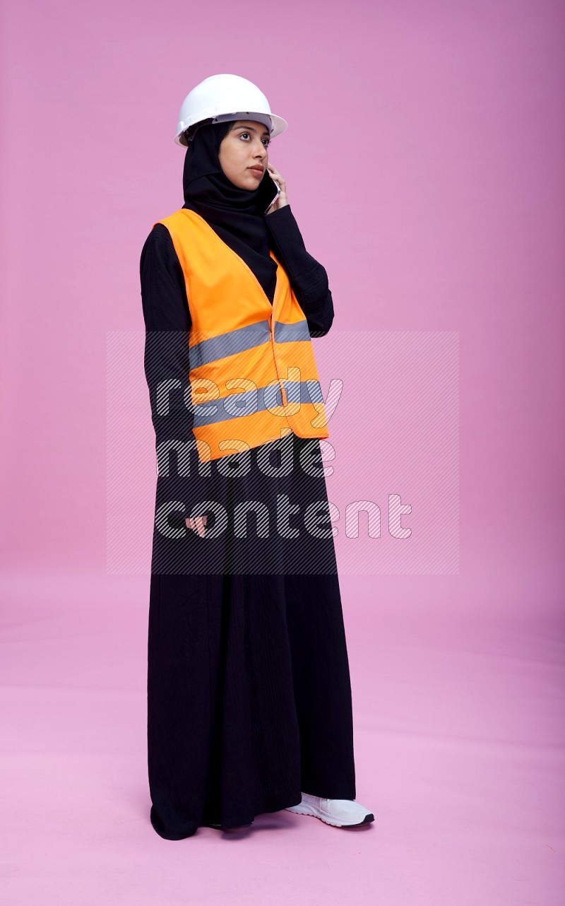 Saudi woman wearing Abaya with engineer vest and helmet standing talking on phone on pink background