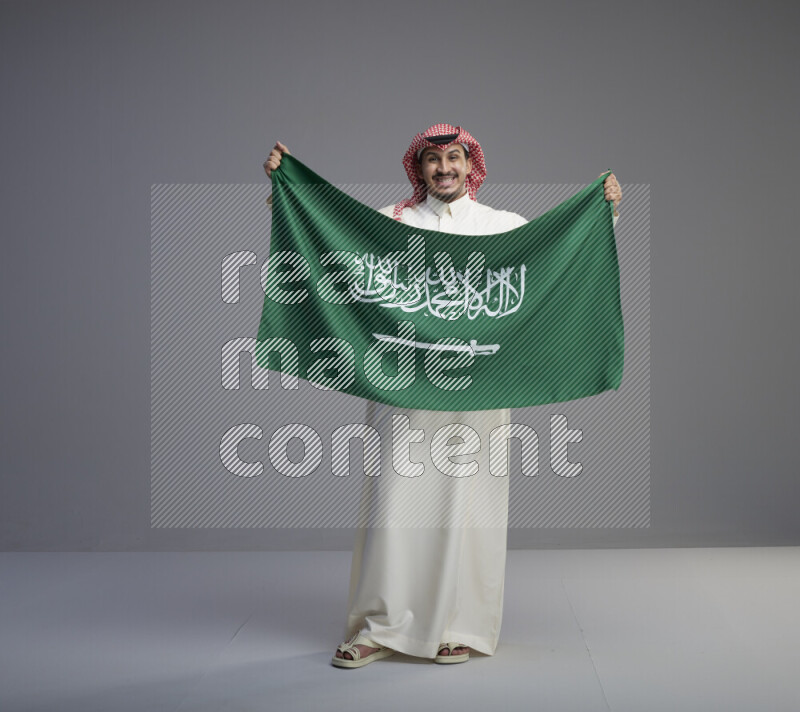A saudi man standing wearing thob and red shomag holding big saudi flag on gray background