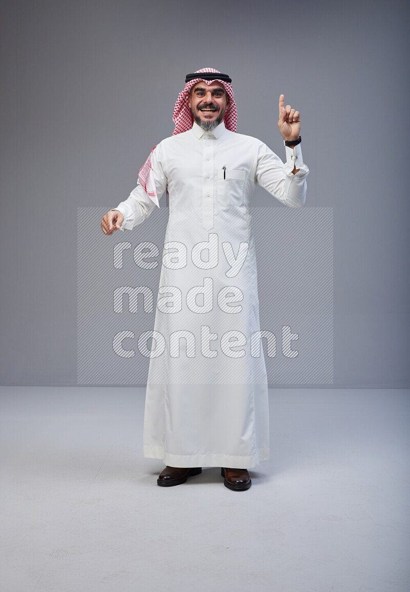 Saudi man Wearing Thob and red white Shomag standing interacting with the camera on Gray background
