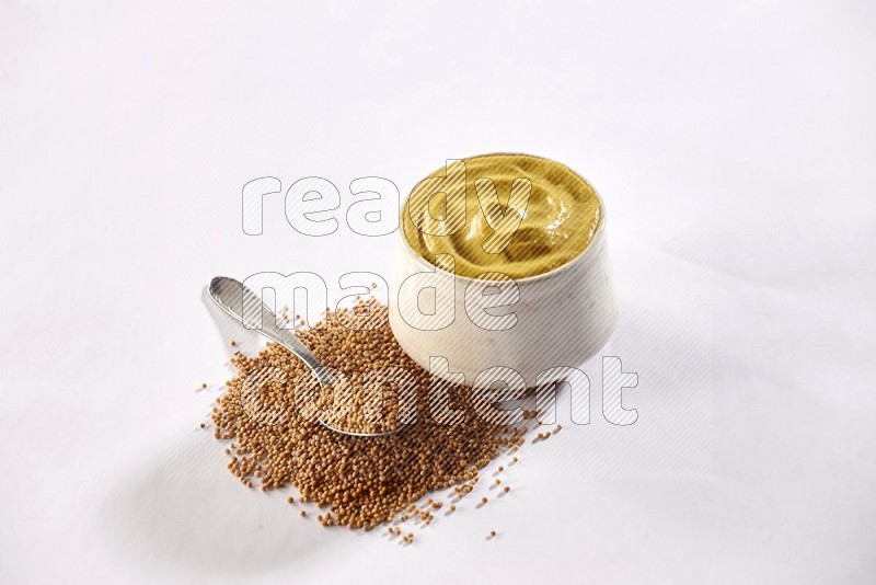 A beige pottery bowl full of mustard paste with mustard seeds underneath with a metal spoon on white flooring in different angles