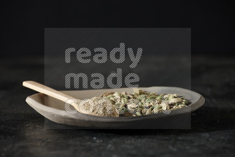 صحن ممتلئ بحبوب الهيل وملعقة خشبية ممتلئة ببودرة الهيل داخل الصحن على خلفية سوداء