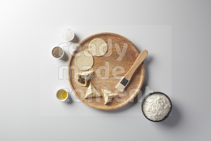 two closed sambosas and one open sambosa filled with meat while flour, salt, black pepper and oil with oil brush aside in a wooden dish on a white background