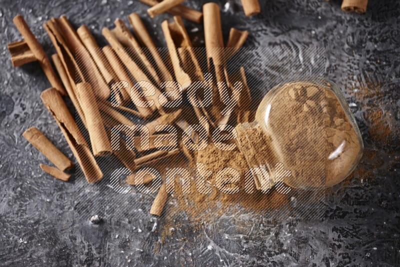 Herbal glass jar full cinnamon powder flipped and a metal spoon full of powder surrounded by cinnamon sticks on textured black background in different angles