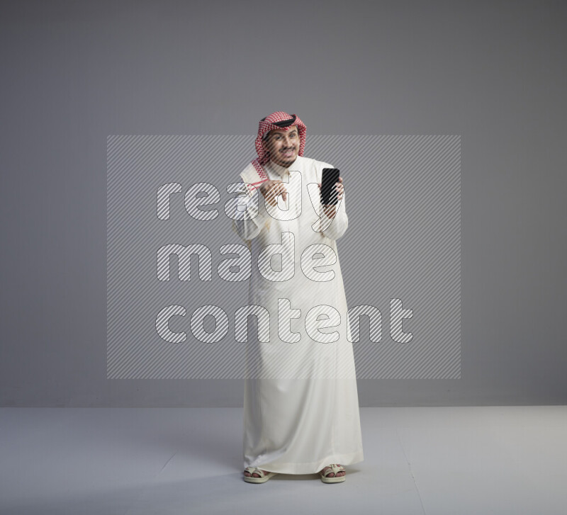 A Saudi man standing wearing thob and red shomag showing phone to camera on gray background