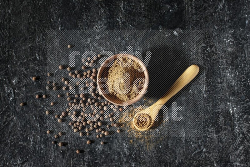 وعاء وملعقة خشبيان ممتلئان ببودرة بهار الفلفل الحلو وحبوبه متناثرة على خلفية سوداء