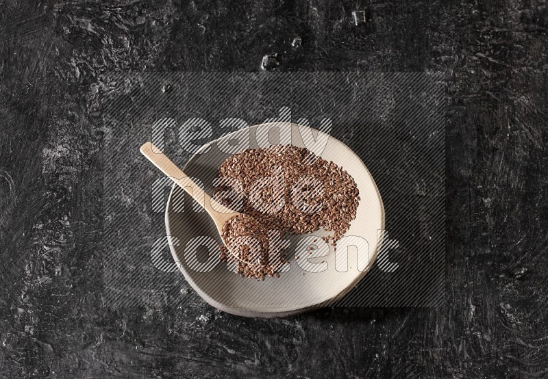 A multicolored pottery plate full of flaxseeds and wooden spoon full of seeds on a textured black flooring
