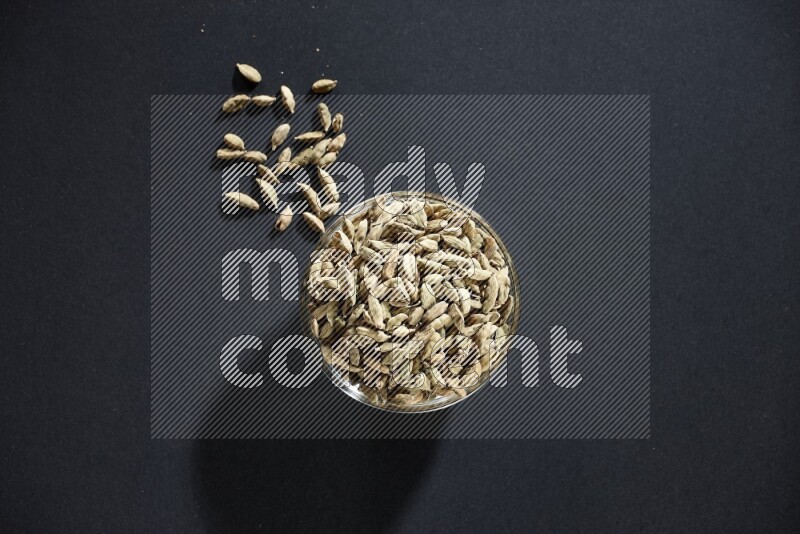 A glass bowl full of cardamom and more seeds spreaded beneath the bowl on black flooring